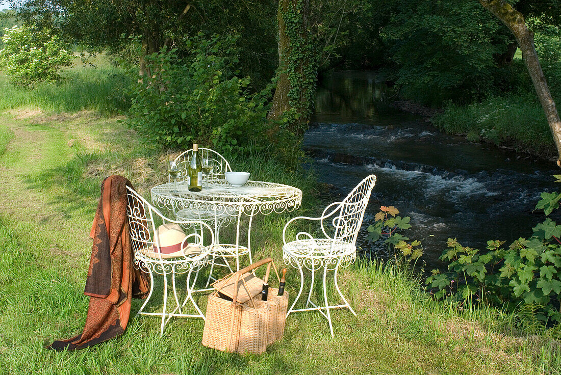 Tisch und Stühle am Flussufer mit einer Auswahl an Wein in einem Korb