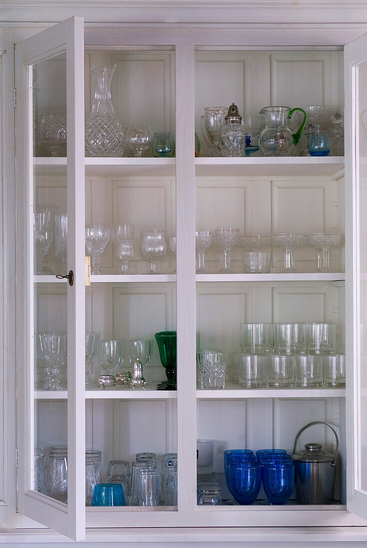 Weiß gestrichene Kommode mit offenen Türen, die eine Sammlung von Glaswaren auf Regalen präsentiert