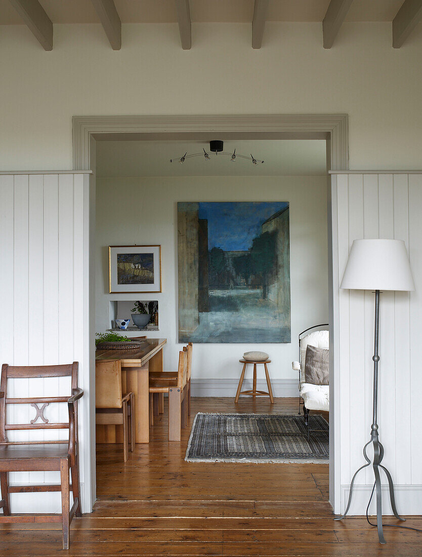 View through to huge abstract painting in deep slate sand and ochre hanging in the dining area along side an upholstered campaign chair and shaker style milking stool with Iron work floorlamp in open plan living area