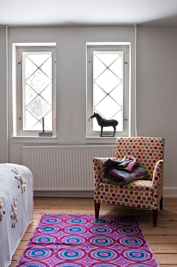Gefaltete Steppdecke auf einem gepolsterten Stuhl mit bunt gemustertem Teppich in einem modernen Schlafzimmer in Odense Dänemark