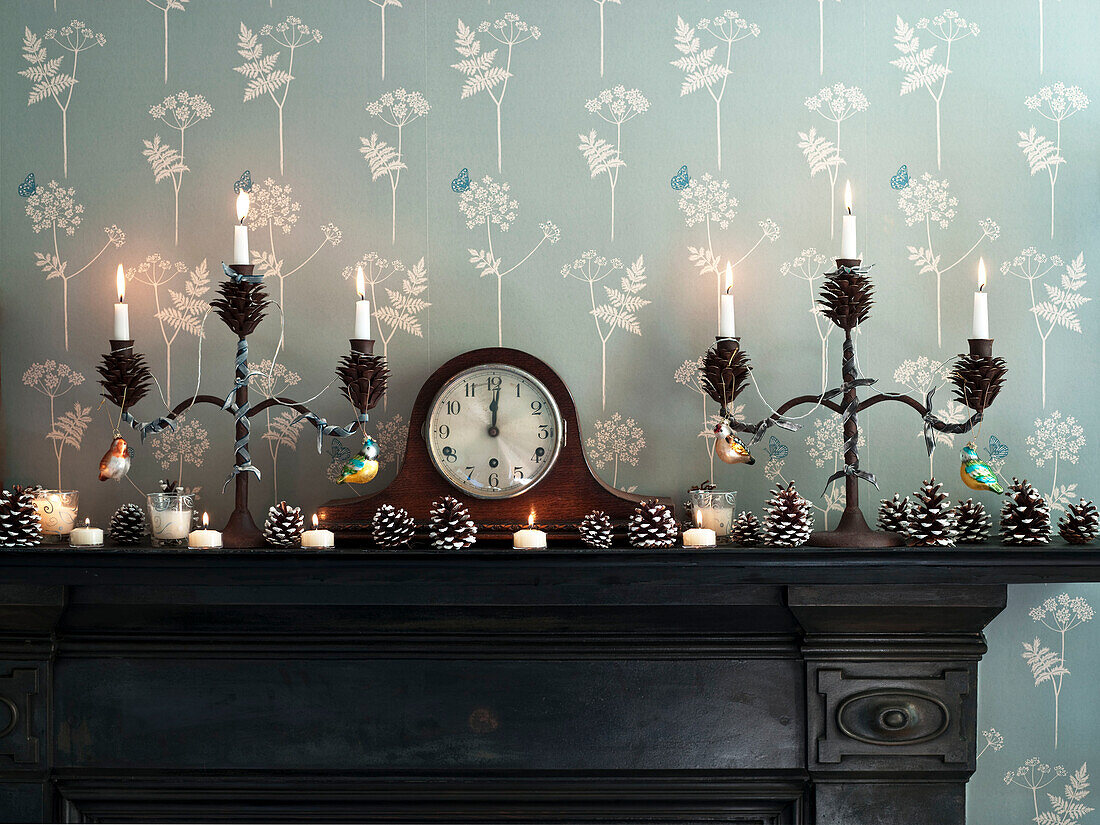 Lit candles with clock and pinecones on fireplace with botanic wallpaper