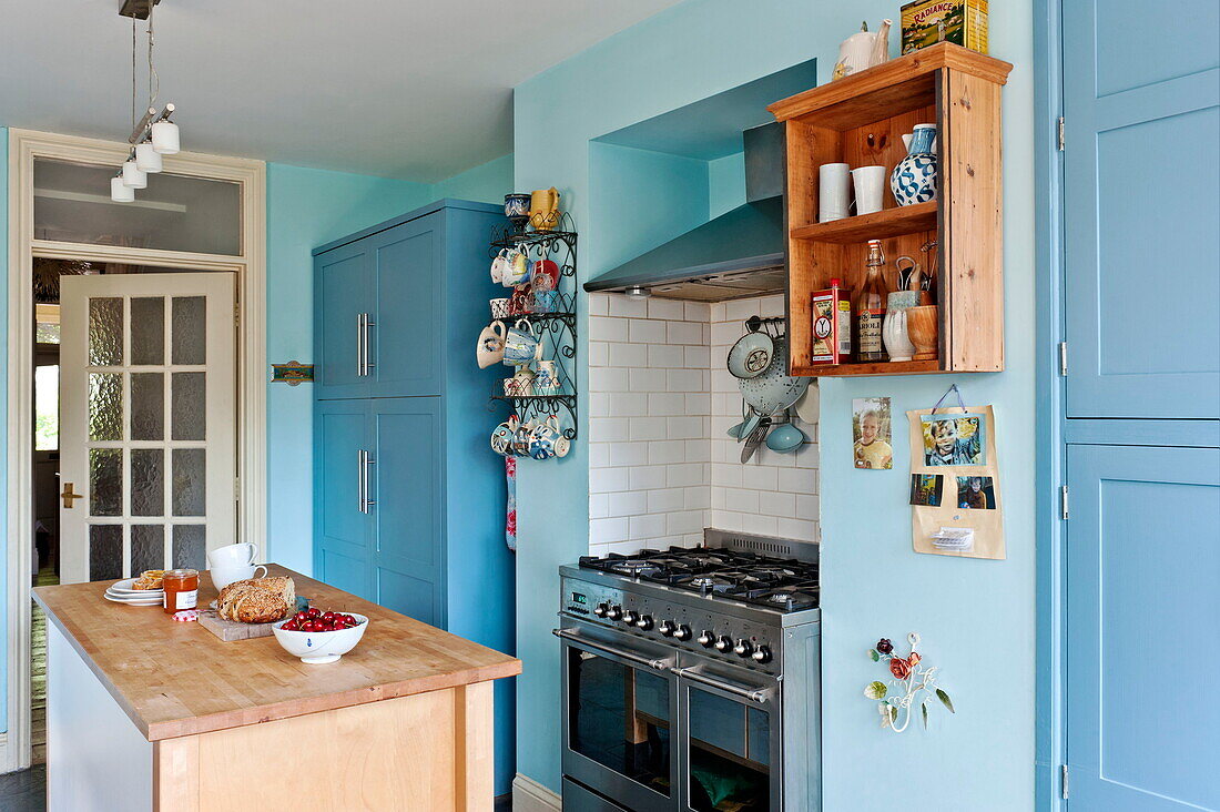 Wooden island unit with wall mounted shelving in blue kitchen of Bovey Tracey family home, Devon, England, UK
