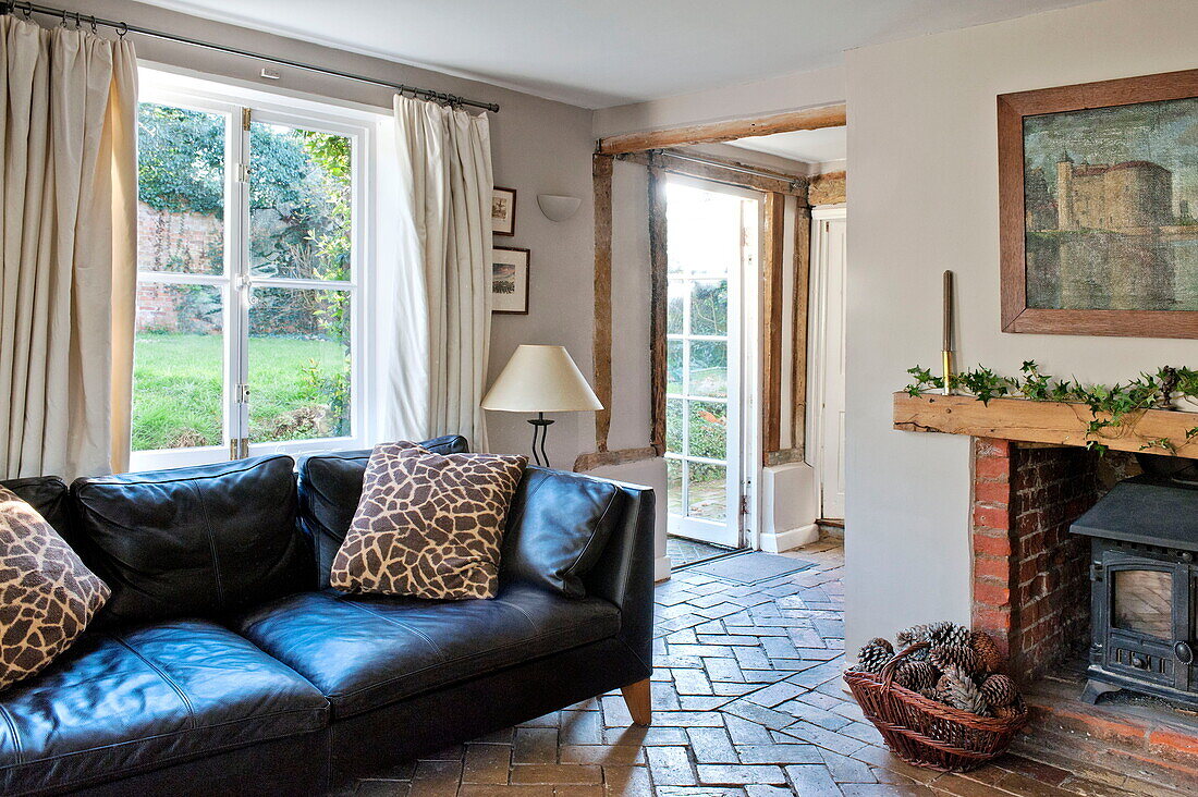 Black leather sofa with wood burning stove in brick floored living room of Suffolk farmhouse, England, UK