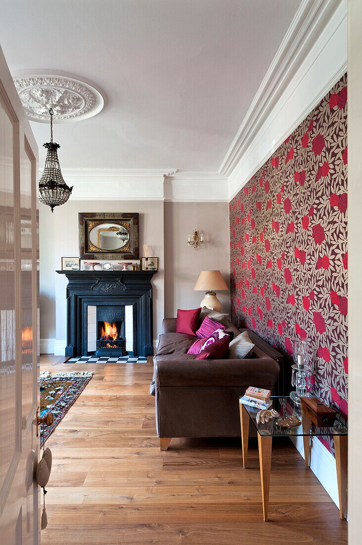 Gemusterte Tapete über braunem Sofa mit beleuchtetem Kamin im Wohnzimmer eines Hauses in Middlesex, London, England, UK