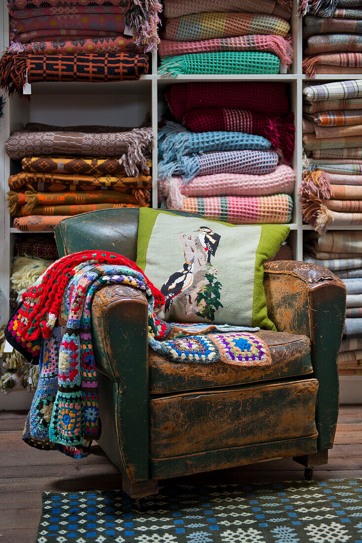 Gehäkelte Decken und Kissen auf einem abgenutzten Ledersessel mit Regalen voller walisischer Decken in einem Geschäft in Tregaon, Wales UK