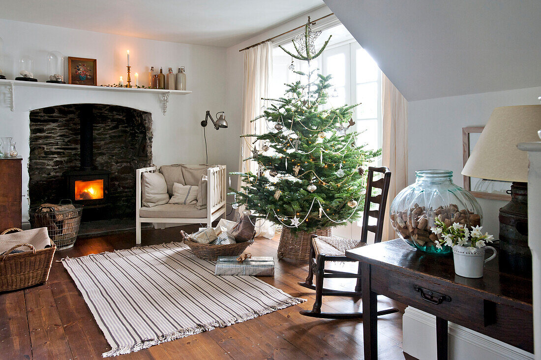 Schaukelstuhl neben dem Weihnachtsbaum mit beleuchtetem Holzofen in einem Haus in Crantock, Cornwall, England UK