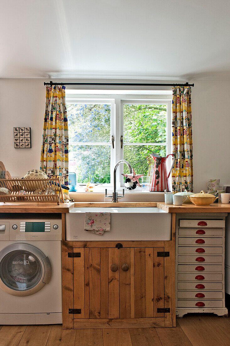 Butler sink and wooden cupboard with washing machine below kitchen window of Cambridge cottage England UK