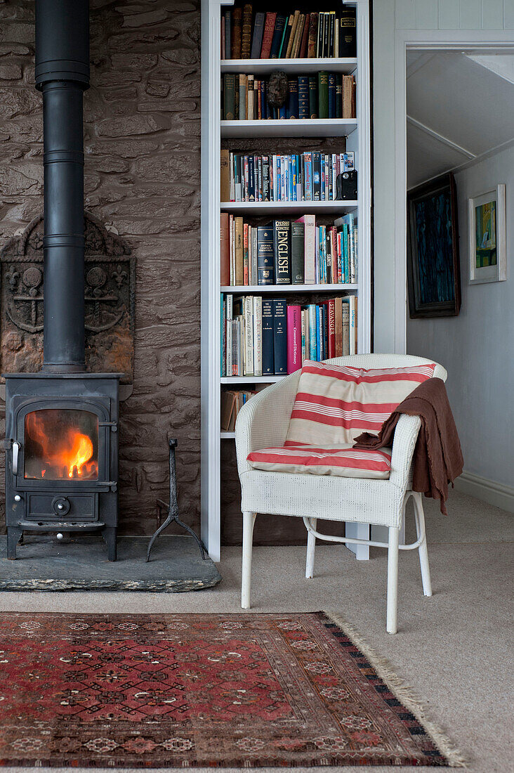 Bookshelf and lit burning stove with chair in beach house Cornwall England UK
