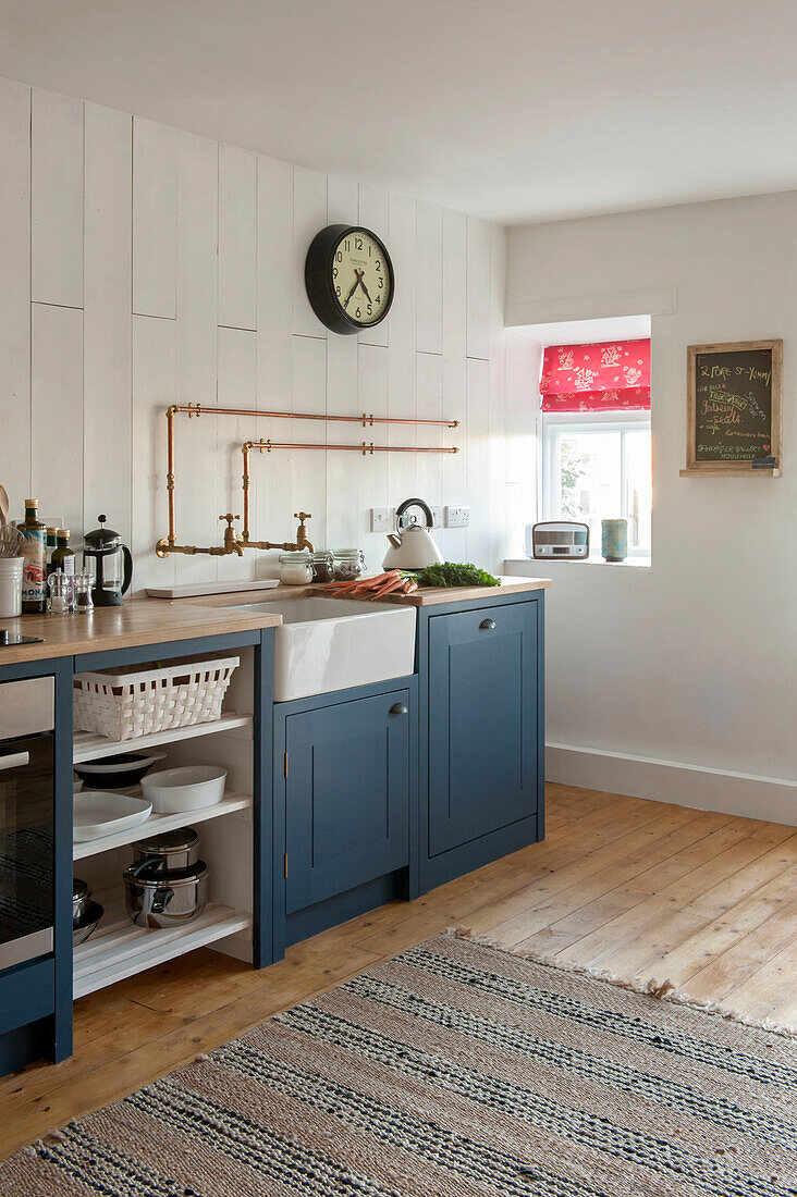 Messing-Wasserrohre und -Uhr über Butler-Spüle mit blauen Einbauschränken in getäfelter Küche in Cornwall UK