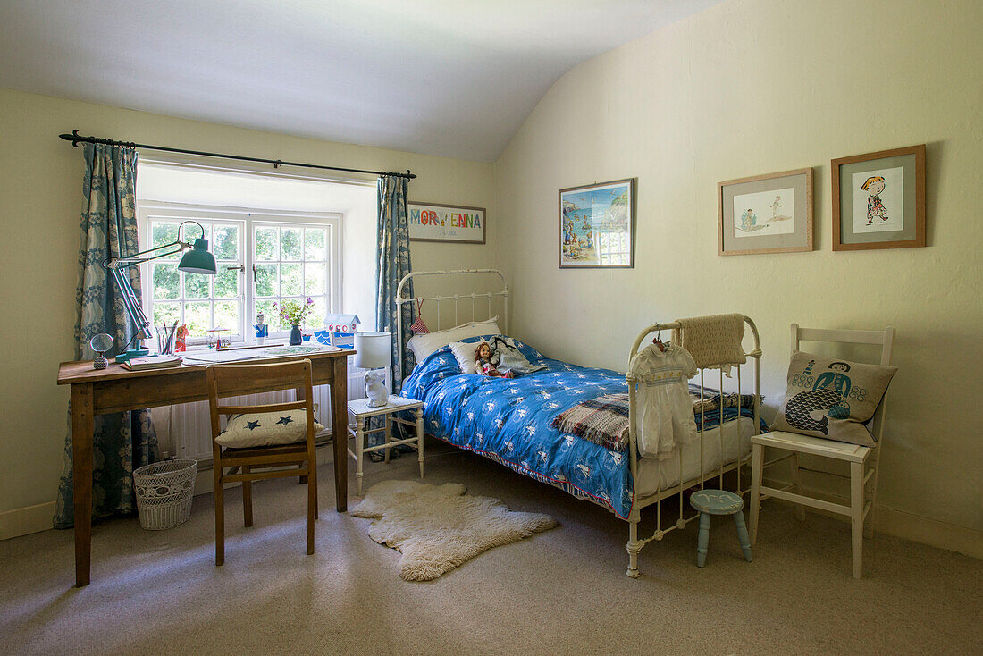 Blaue Bettdecke auf metallgerahmtem Einzelbett mit Schreibtisch und Stuhl am Fenster in einem Bauernhaus in Helston, Cornwall, Großbritannien
