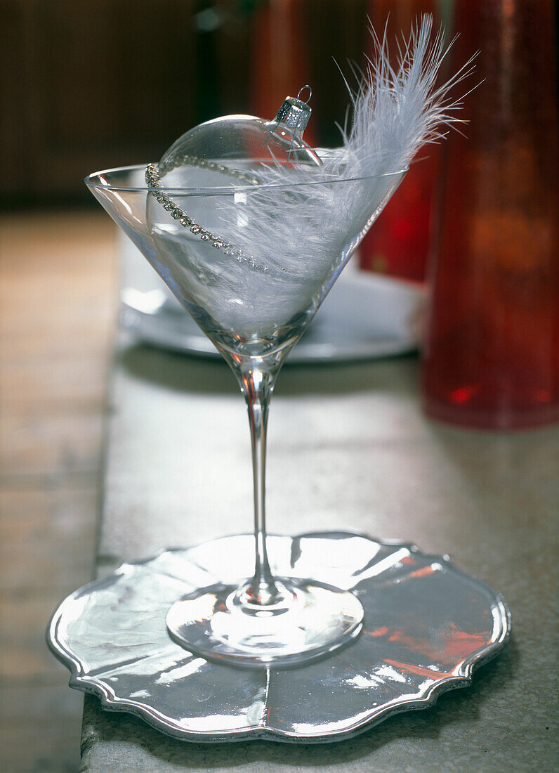 Martini-Glas mit Christbaumkugel und Feder auf Tischplatte