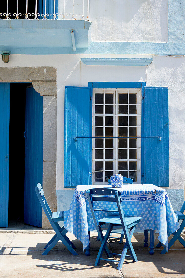 Blaue Klappstühle am Tisch vor einer sonnenbeschienenen griechischen Villa