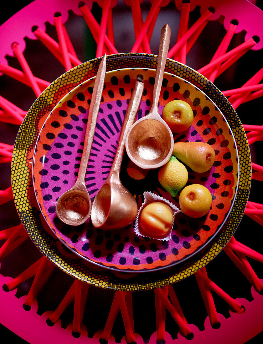 Goldfarbene Metalllöffel und künstliche Früchte auf einem bunt gemusterten Teller