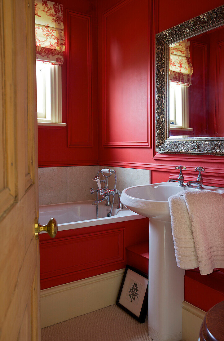 Blick durch eine Holztür auf ein rotes Badezimmer mit weißem Keramikwaschbecken im Badezimmer eines Bauernhauses in Etchingham, East Sussex, England, UK