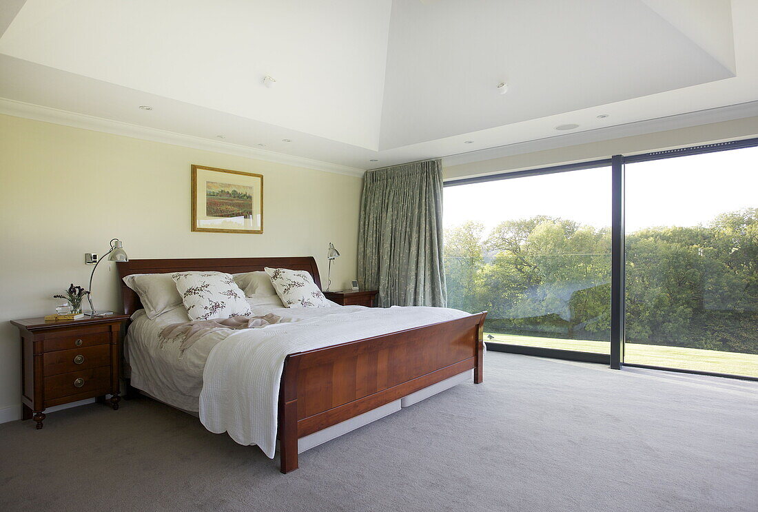Polished wooden double bed in room with full length windows to garden, contemporary farmhouse, Nuthurst, West Sussex, England, UK