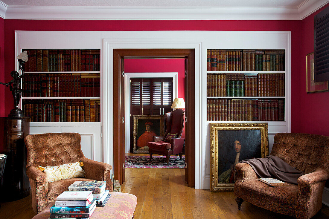Pair of upholstered chairs in library with hardbacked books and gilt framed oil painting in Kent home England UK