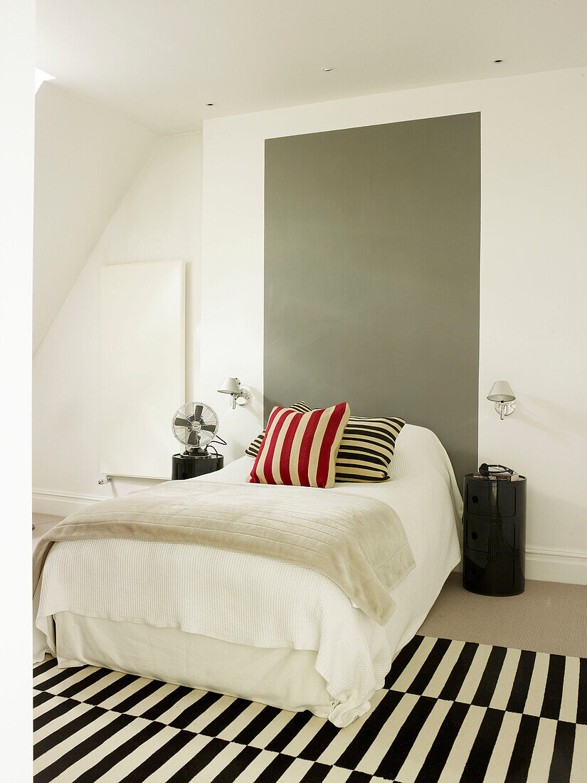 Graues Paneel am Kopfende des Bettes mit rot gestreiftem Kissen und schwarz-weißem Teppich in einem Haus in London, UK