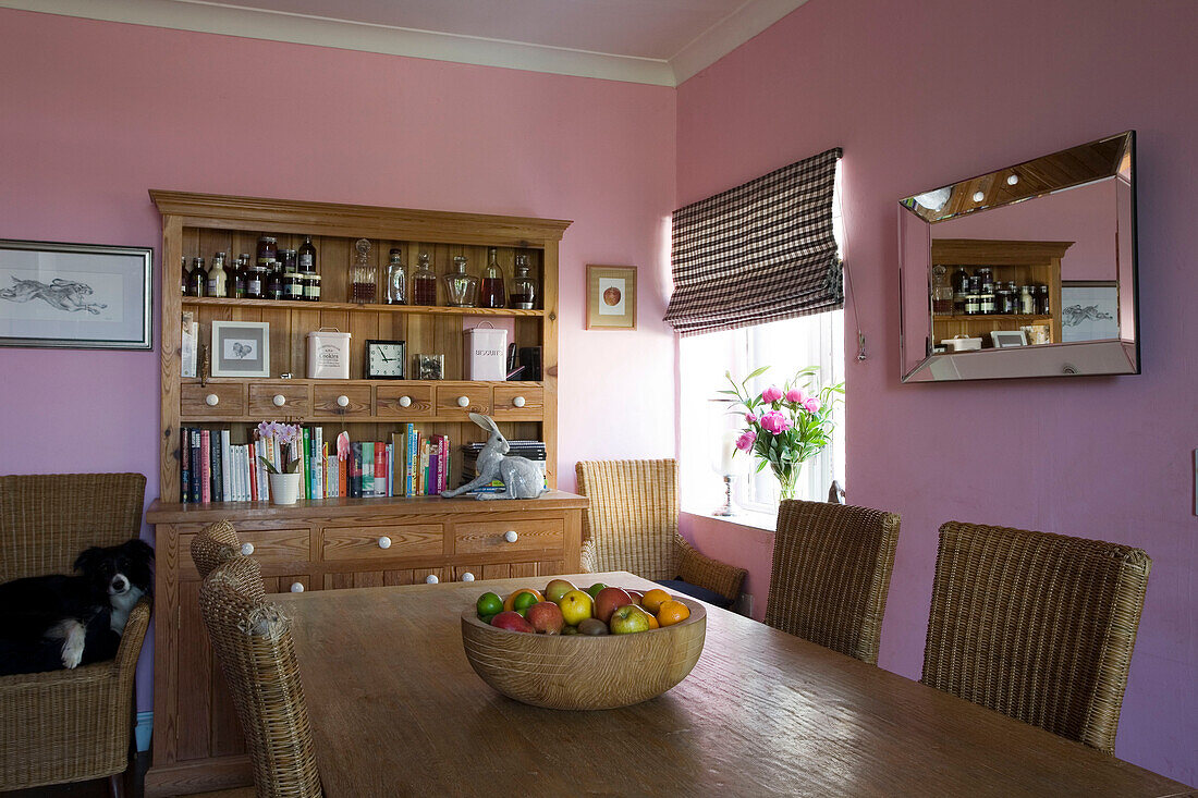 Wooden fruitbowl on table with dresser and dog sleeping on rattan chair in pink Suffolk dining room UK