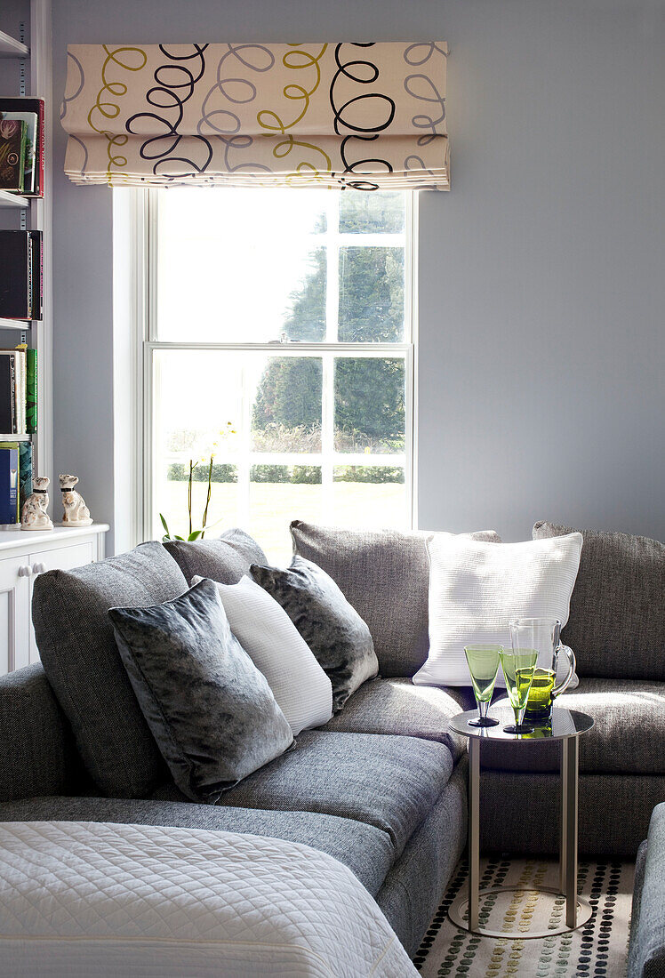 Roman blinds at sunlit window of Suffolk living room with grey sofas UK