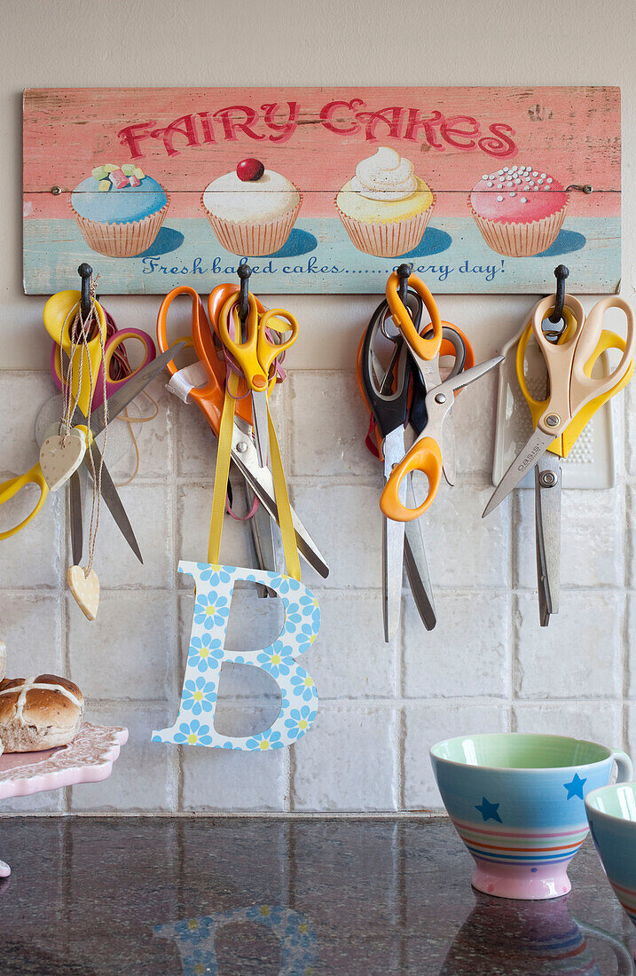 Scissors hang on kitchen hooks with teacups and tiled splashback in Berkshire home, England, UK