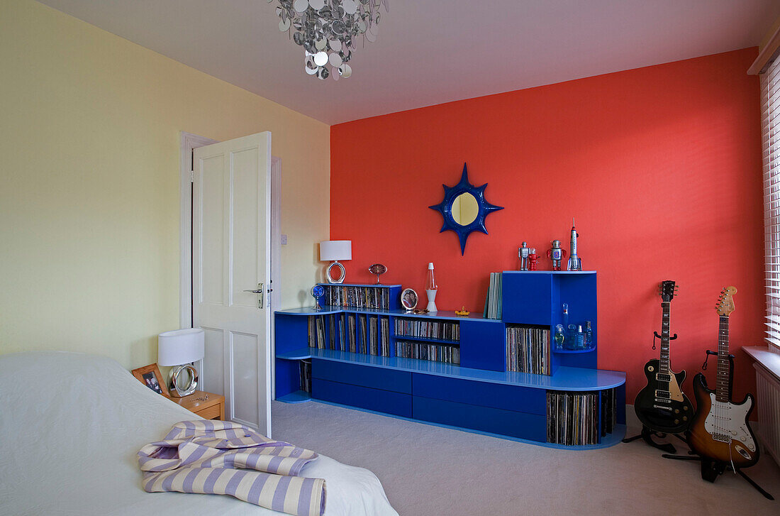 Bright blue storage unit and electric guitars set against red wall in bedroom of retro styled East Sussex home, England, UK