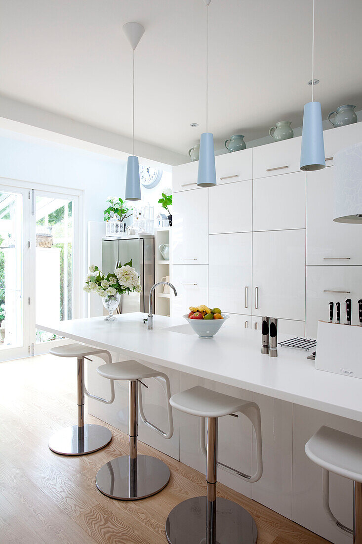 Bar stools at breakfast bar in white fitted kitchen with light blue pendants in contemporary London home, UK