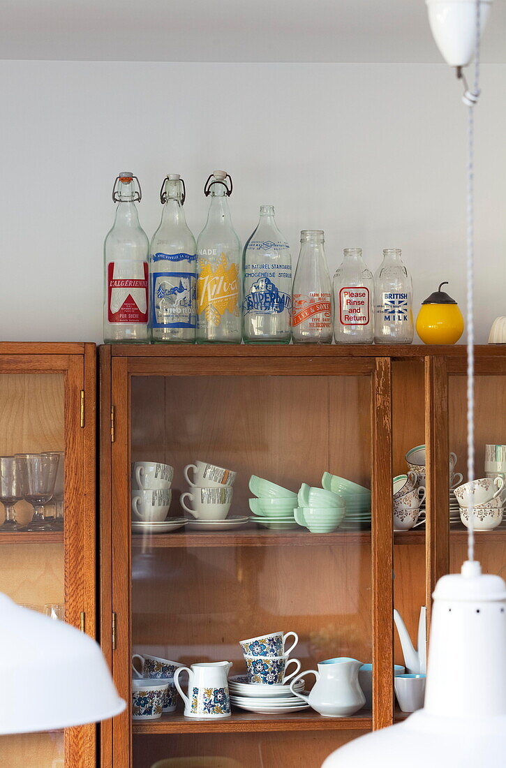 Teetassen und Geschirr mit alten Flaschen in einem hölzernen Küchenschrank mit Glasfront in einem Familienhaus in Kent (England)