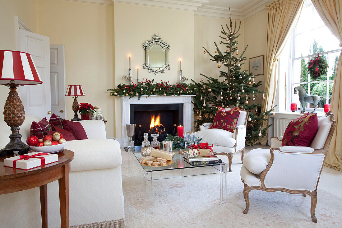 Ein Paar passender Sessel und ein beleuchtetes Feuer mit Weihnachtsbaum im Wohnzimmer in West Sussex, England, Vereinigtes Königreich
