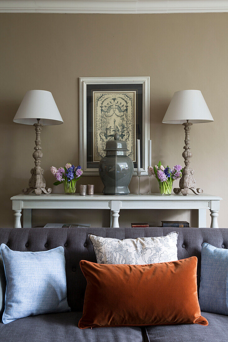 Passende Lampen auf einer Konsole hinter einem grauen, geknöpften Sofa mit einer Vielzahl von Kissen in einem Haus in London, England, Vereinigtes Königreich
