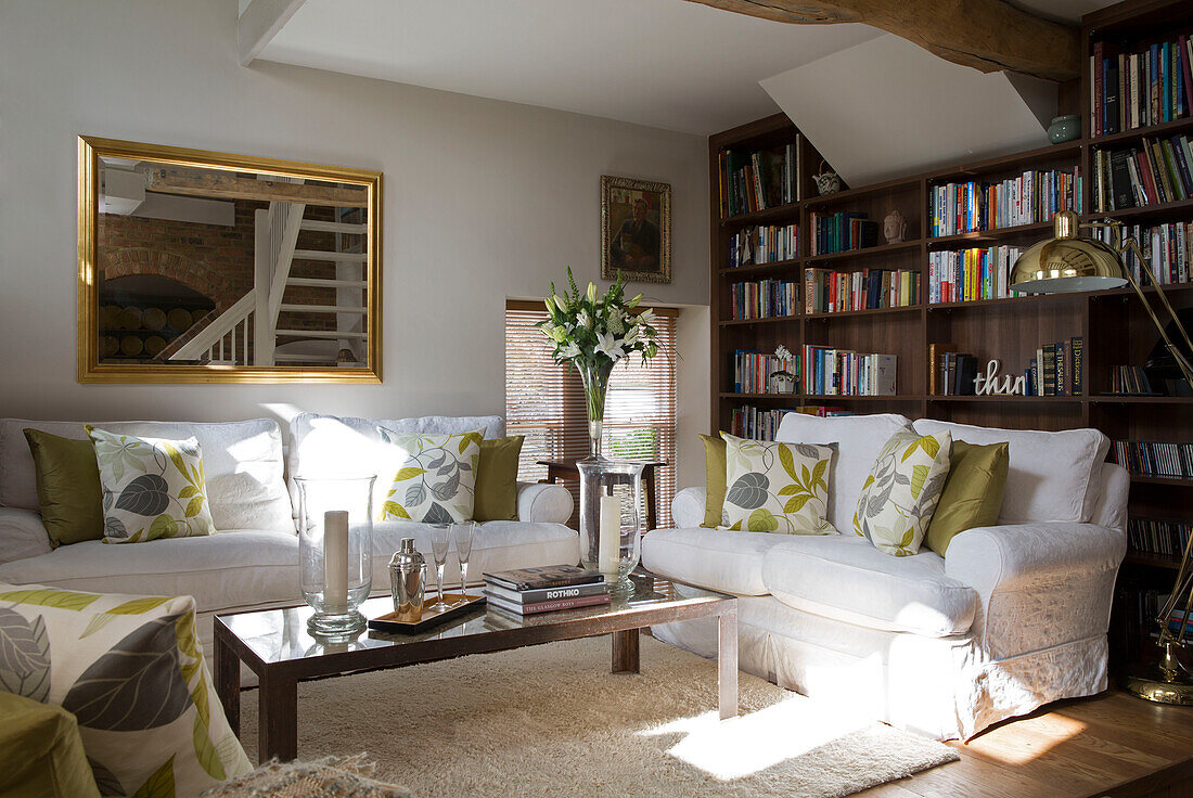 Blattgemusterte Kissen auf weißen Sofas mit Bücherregalen und großem, goldgerahmten Spiegel im Wohnzimmer eines britischen Bauernhauses
