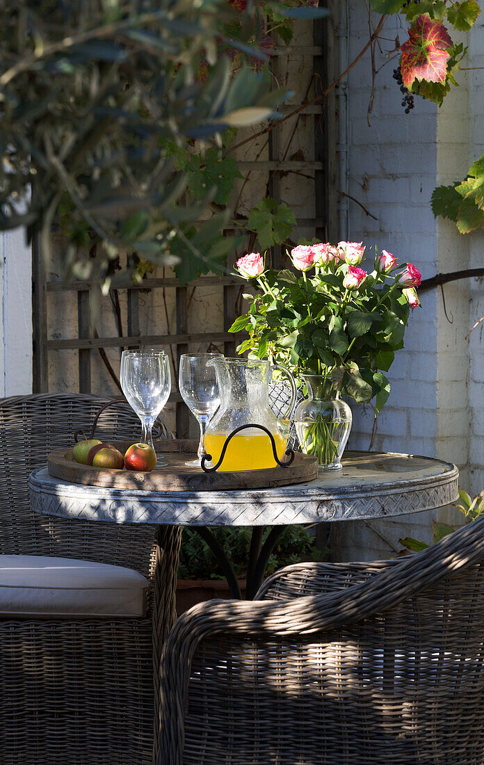 Krug mit Saft und Äpfeln mit geschnittenen Rosen auf Tisch mit Korbsesseln im Innenhof eines Hauses in Berkshire, England, UK