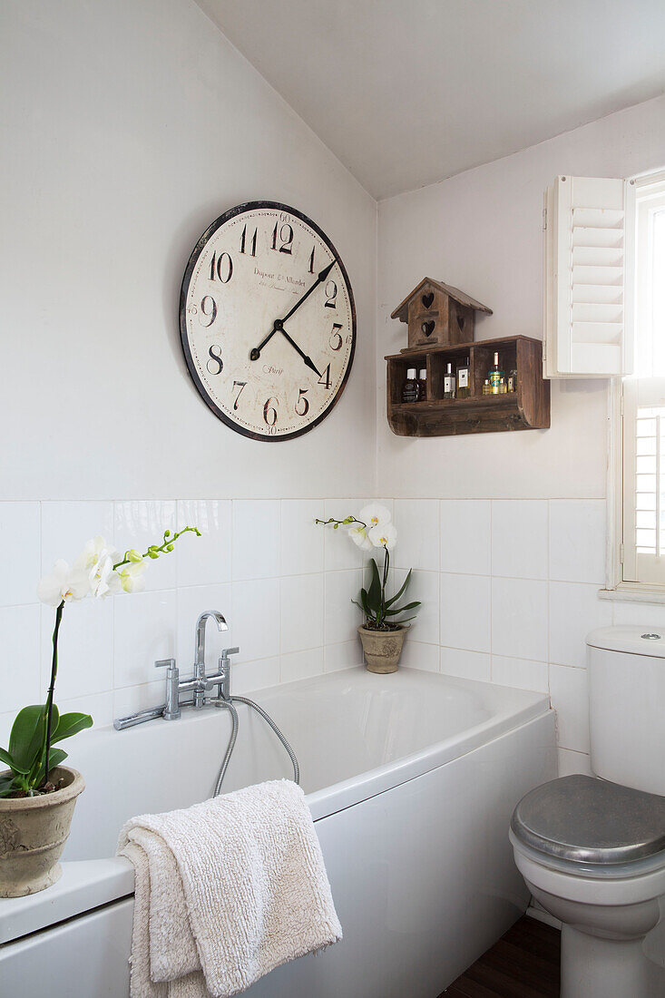Große Uhr und Regal über Orchideen auf weißer Badewanne mit Mischbatterie in einem Haus in Berkshire, England, UK