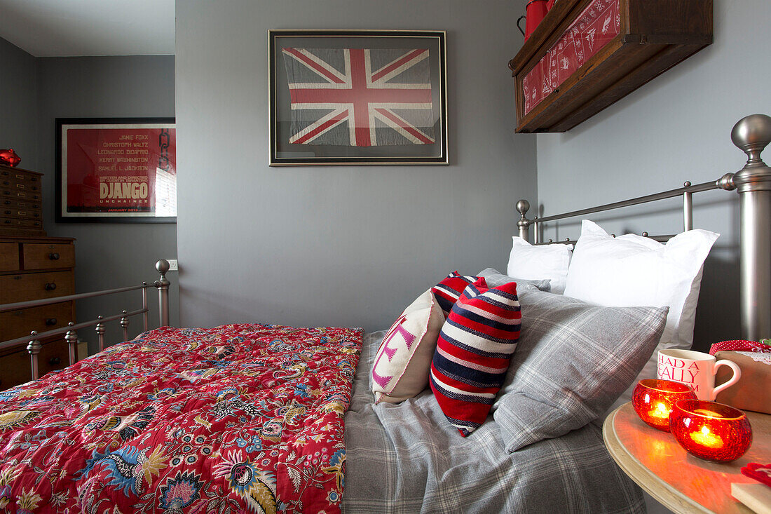 Gestreifte Kissen mit gemusterter roter Steppdecke auf silbernem Schmiedeeisenbett in grauem Schlafzimmer mit Union Jack in Surrey Schlafzimmer England UK