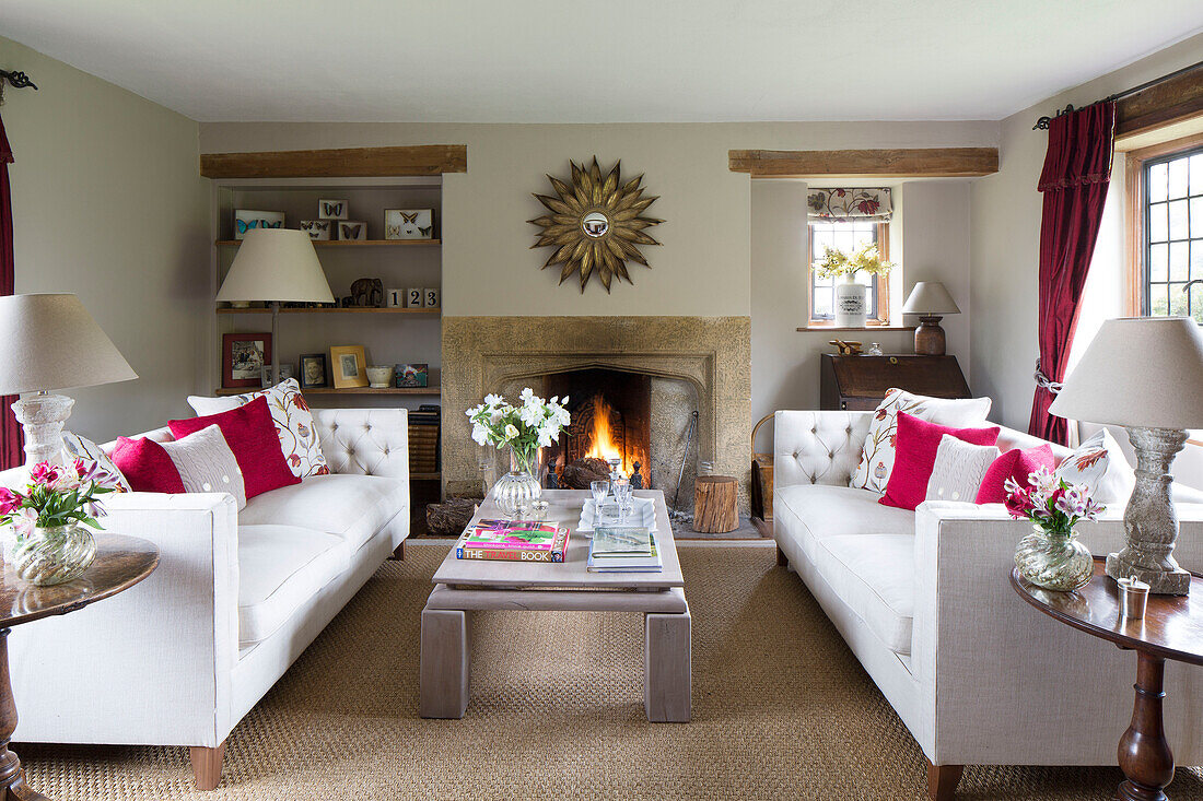 Pair of white sofas with low table and sunburst mirror above lit fire in Sussex farmhouse   England   UK