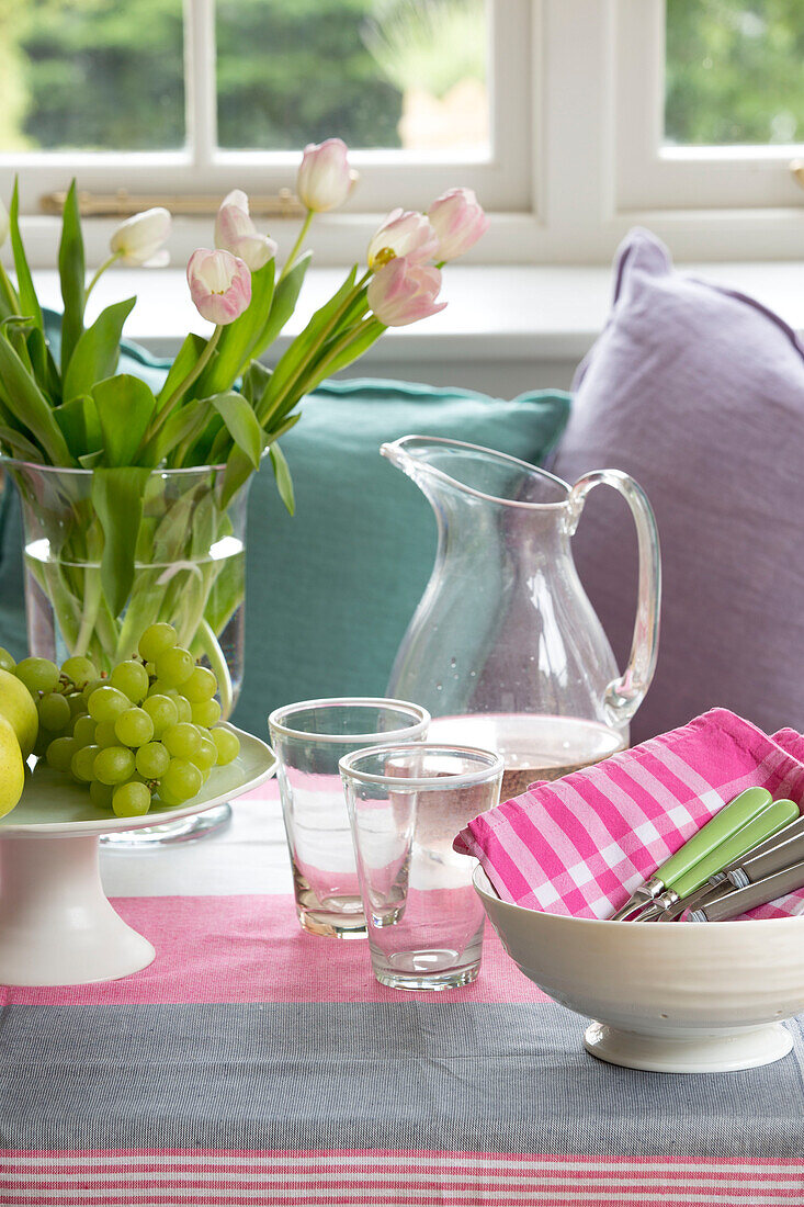Trinkwasser und Gläser mit geschnittenen Tulpen und Weintrauben auf gestreifter Tischdecke in einem Haus in Gloucestershire, England, Vereinigtes Königreich