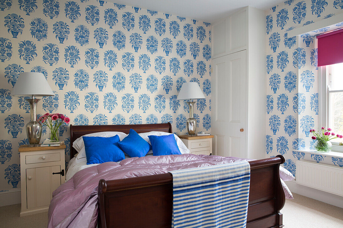 Blau gemusterte Tapete über einem Holzbett mit passenden Nachttischlampen in einer Wohnung in Gloucestershire, England, UK