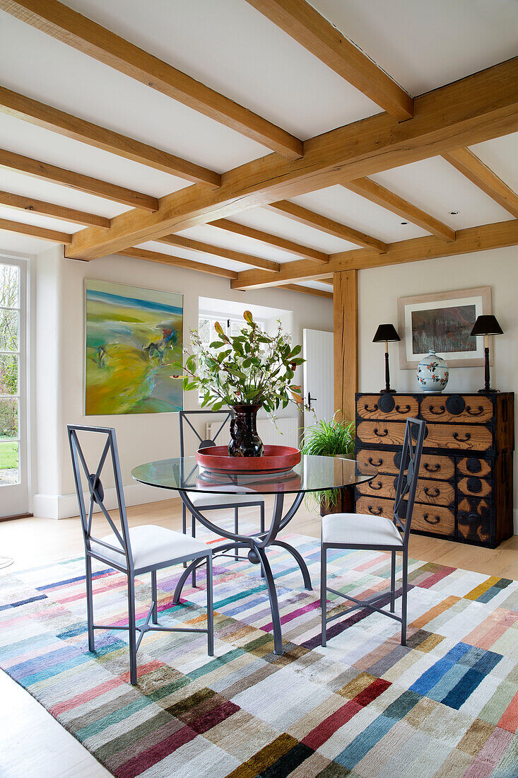 Glass topped table with metal chairs and patterned rug under beamed ceiling in Gloucestershire farmhouse England UK