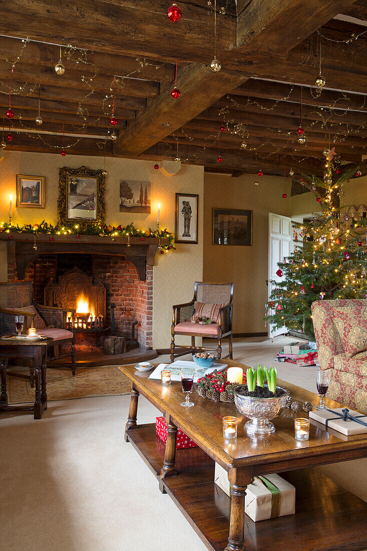 Beleuchtetes Feuer und Weihnachtsbaum mit Weihnachtsdekoration in Deckenbalken im Wohnzimmer eines Bauernhauses in Hampshire England UK