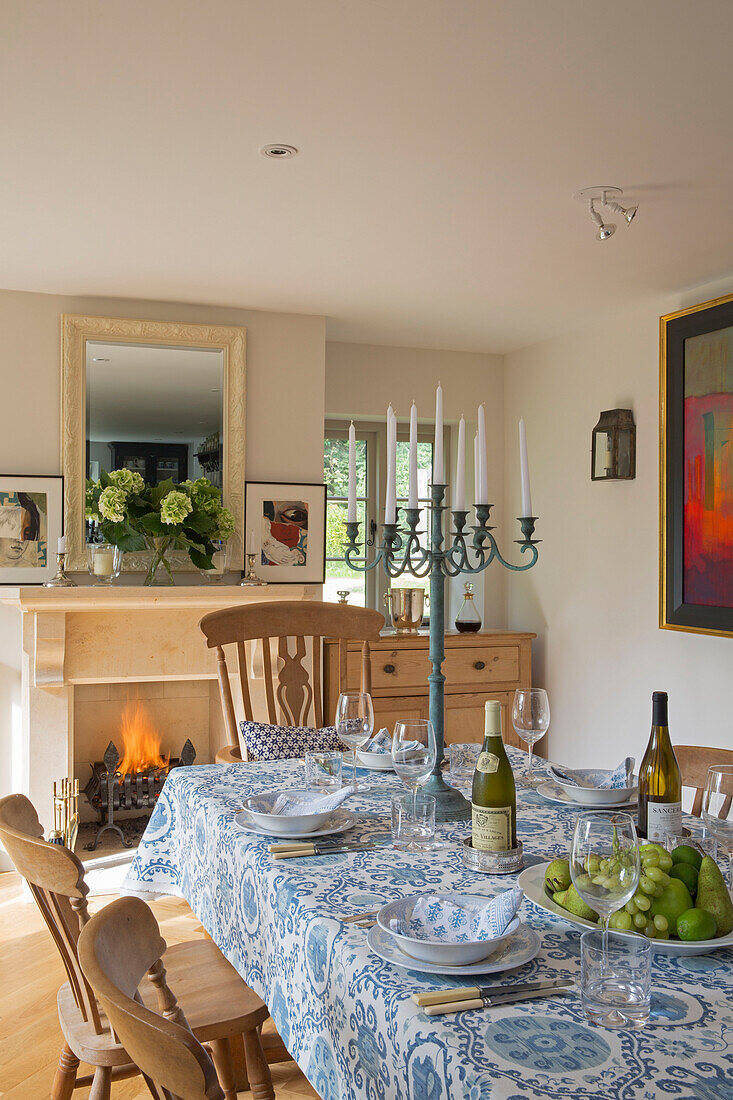 Kerzenständer und Weinflaschen auf blauem und weißem Tischtuch mit beleuchtetem Feuer im Esszimmer eines Landhauses in Gloucestershire UK