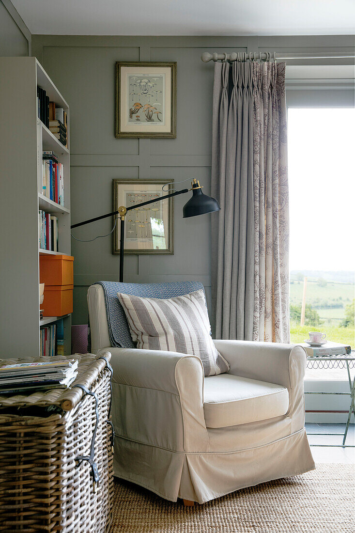 Slip covered armchair with bookshelf and basket at window in Somerset farmhouse UK