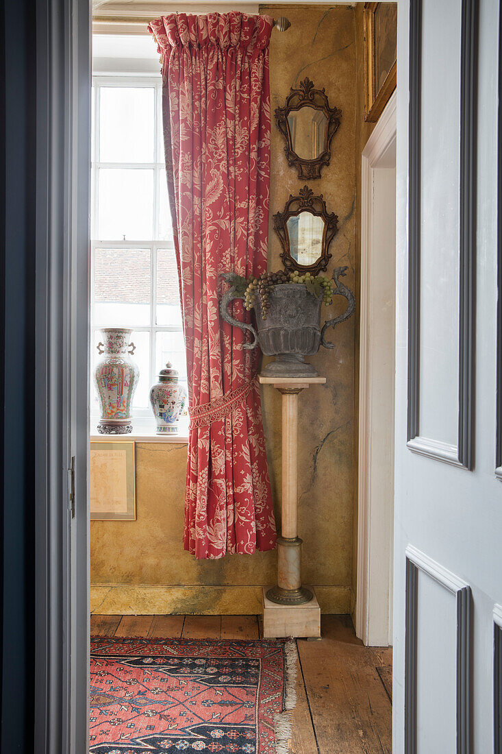 Urn on pedestal with antique mirrors and red curtains at window in Sussex home