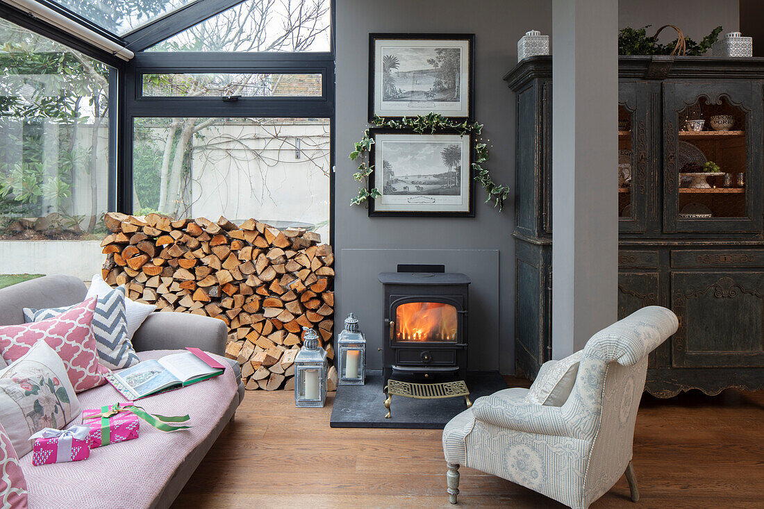 Armchair and sofa with stacked wood and lit fire in conservatory extension of London villa UK
