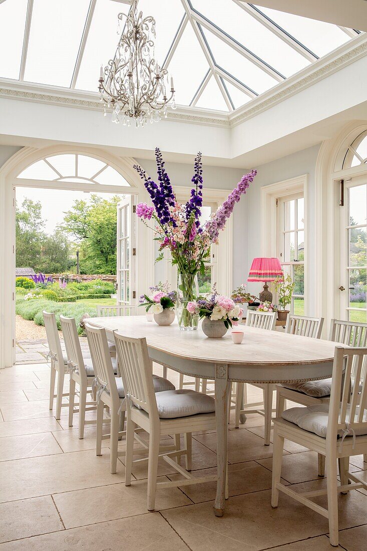 Elegant light filled conservatory room with dining table and chairs festooned with fresh cut flowers with open doors leading on to the garden in Hampshire UK