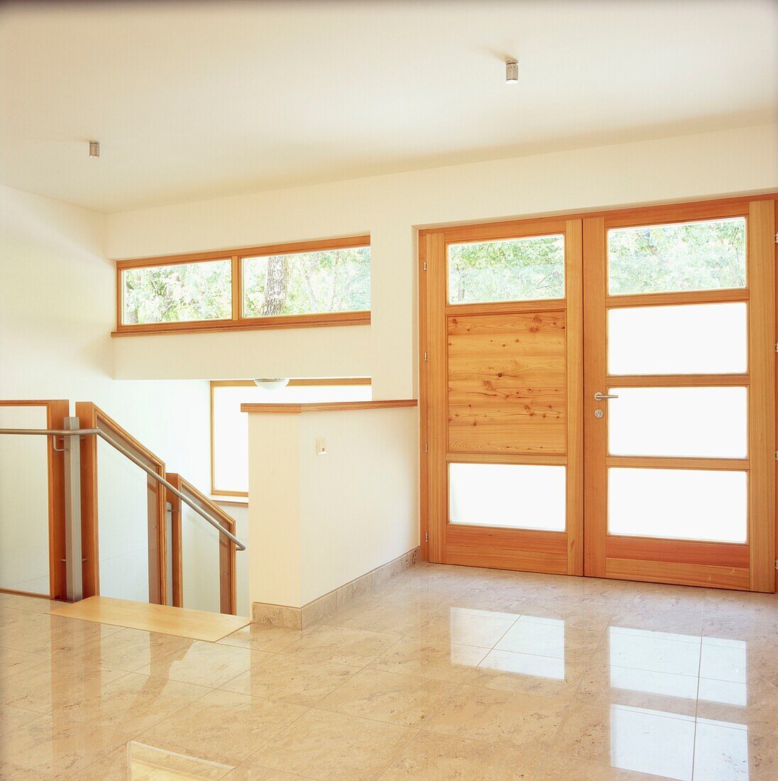 Large contemporary empty entrance hall with windows double wood and glass front door white walls and a marble tiled floor