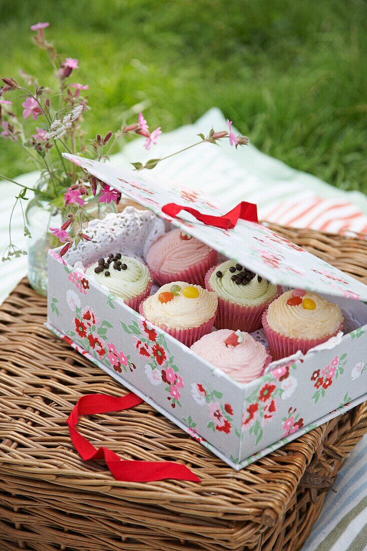 Schachtel mit hübschen, glasierten Cupcakes in einer mit Blumen bedeckten Tortenschachtel auf einem Weidenkorb mit einem Gefäß mit Wildblumen