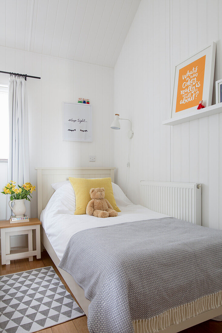 Einzelbett mit Teddybär und Nachttisch mit Blumen in weißem Zimmer