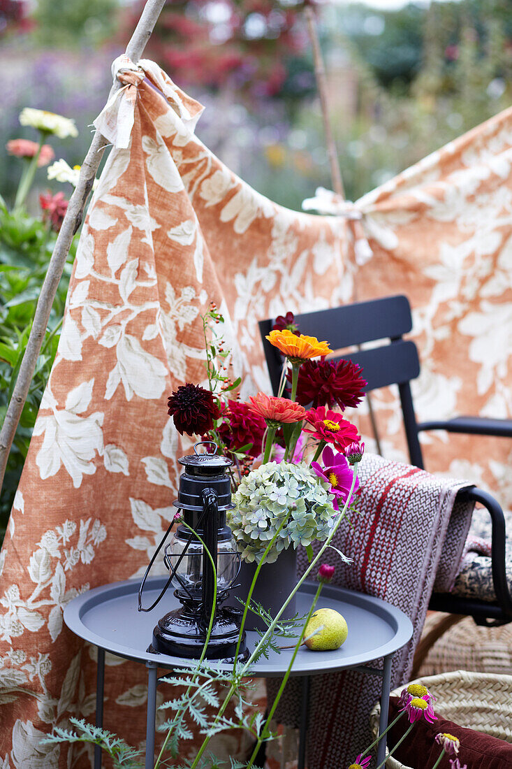 Blumendekoration und Laterne auf Beistelltisch im Garten, gemustertes Tuch als Abtrennung