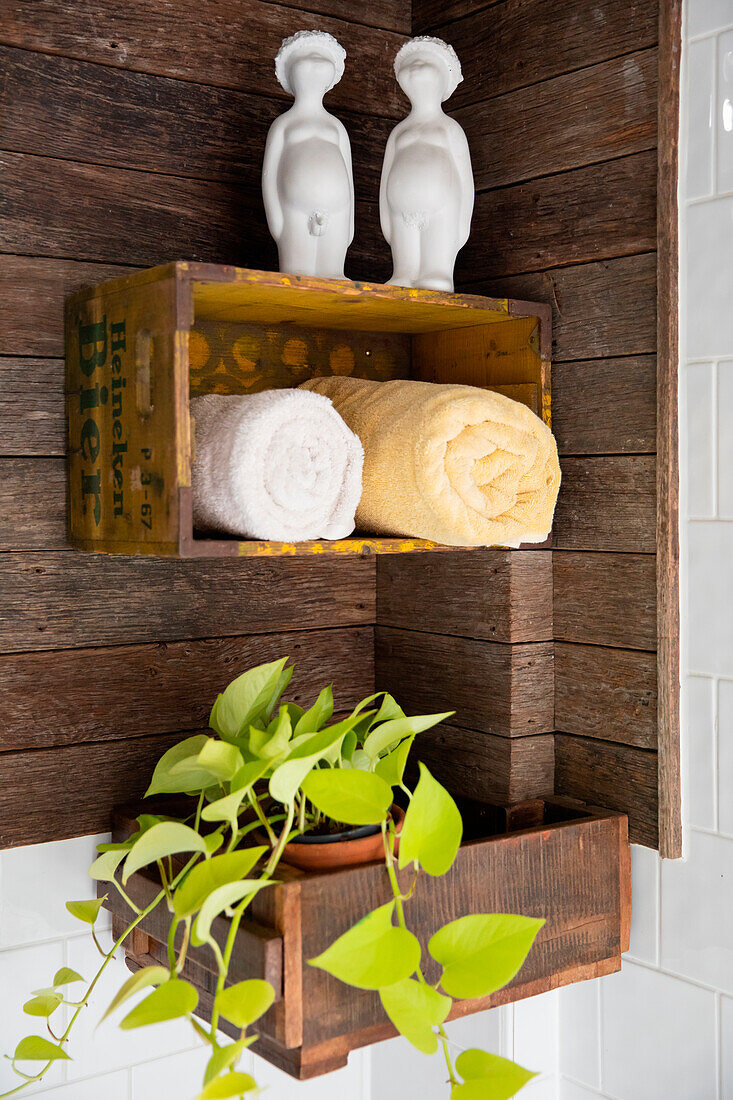 Holzkiste mit Skulpturen als Handtuchablage an Bretterwand, darunter Zimmerpflanze