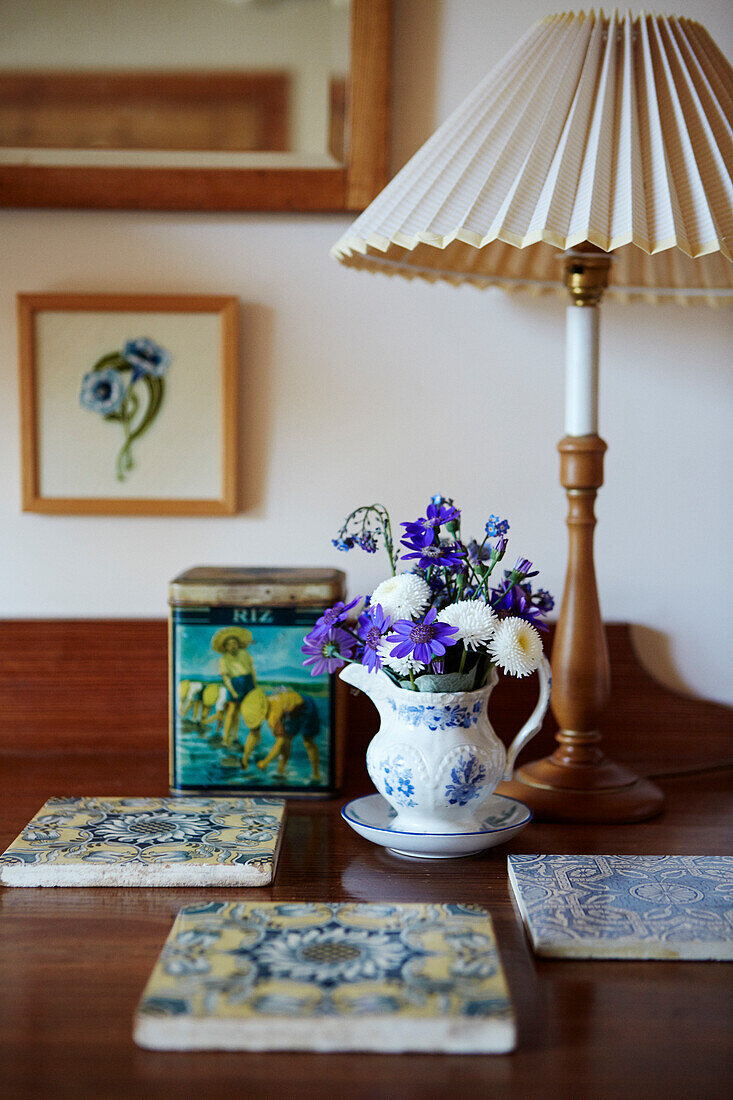 Tile coasters, posy of flowers in jug and table lamp on wooden table