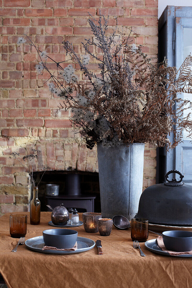 Gedeckter Tisch mit Trockenblumen in Metallvase vor Ziegelwand